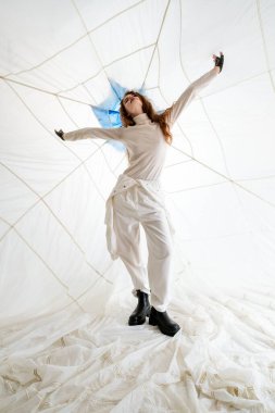 Uzun kahverengi saçlı genç bir kadın ağ tavanlı beyaz bir odada duruyor. O, beyaz elbise ve siyah botlar giyiyor. Kolları açıkta ve o da tavana bakıyor. Şey...