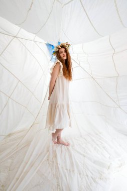Beyaz bir elbise ve çiçek tacı giyen bir kadın büyük bir kumaş yapının üzerinde duruyor. Kumaş beyazdır ve hafiflik ve hava hissi yaratır. # Kızıl saçlı ve çıplak ayaklı kadınlar #