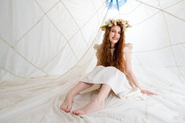 Uzun kızıl saçlı ve taç takmış genç bir kadın büyük, beyaz bir çadırın içinde bir yatakta oturuyor. O, beyaz bir elbise giyiyor ve bir gülümsemeyle kameraya bakıyor. Çadır beyaz kumaştan yapılmıştır.