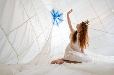 Uzun, kızıl saçlı ve beyaz elbiseli genç bir kadın büyük, beyaz, kumaş bir yapının içinde oturuyor. Kadın kolunu binanın tepesine doğru uzatıyor. Mavi dairesel bir nesnenin olduğu yere.