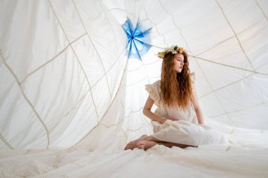 Beyaz bir elbise ve çiçek tacı giyen bir kadın büyük beyaz bir gölgeliğin içinde oturur. Aşağıya bakıyor ve uzun kızıl saçları var. Kubbe beyaz kumaştan yapılmıştır ve üzerinde mavi bir tasarım vardır..