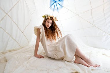 Uzun, dalgalı kızıl saçlı genç bir kadın beyaz bir çadırda otururken beyaz bir elbise ve çiçek tacı giyer. Çadırın üzerinde beyaz, desenli bir kumaş ve mavi bir şemsiye asılı.