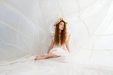 Uzun kızıl saçlı ve çiçek kaplamalı genç bir kadın beyaz kumaş bir yapının üzerinde oturuyor. O, fırfırlı kollu beyaz bir elbise giyiyor. Kumaş yapısının geometrik bir deseni var, çizgileri ve