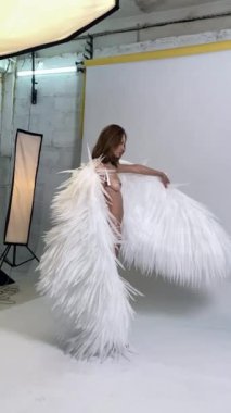 Beyaz bir stüdyoda büyük beyaz tüylü kanatları olan bir kadın poz veriyor. Önünde tutuyor ve nazikçe hareket ettiriyor..