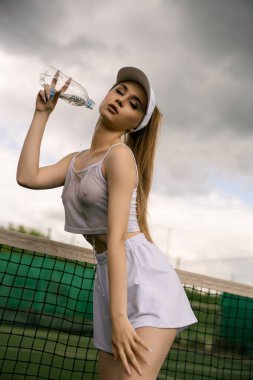 Genç bir bayan sporcu, elinde su şişesiyle tenis kortunda poz veriyor. Dramatik bir gökyüzünün altında spor kıyafetini sergiliyor..