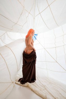 Pembe saçlı bir kadın beyaz kumaş bir yapının içinde kahverengi bir bornoz giyiyor. Yapının dokusu gerilmiş, ağ benzeri bir desen oluşturuyor. Sağ tarafa doğru bakıyor.