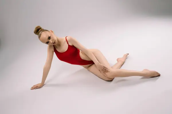 stock image A dancer in a red swimsuit strikes a graceful pose on the studio floor, showcasing flexibility and elegance in soft lighting.