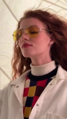 Kızıl saçlı genç bir kadın beyaz ceket ve renkli damalı gömlek giyerken sarı renkli güneş gözlüğü takıyor..