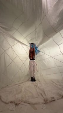 Beyaz pantolonlu ve siyah beyaz gömlekli bir kadın büyük beyaz bir paraşütün içinde dans ediyor..