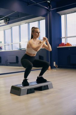 Atletik giyinen bir kadın, iyi ışıklandırılmış bir spor salonundaki bir platforma çömelir. Çevre egzersiz için tasarlanmıştır, arka planda renkli ağırlıklar görünür..