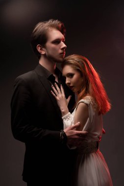 Siyah takım elbiseli kendine güvenen genç adam siyah arka planda duran gelinlik giymiş kadını kucaklıyor.