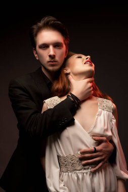 Siyah takım elbiseli kendinden emin genç adam siyah arka planda dururken gelinlik giymiş kadını boğuyor.