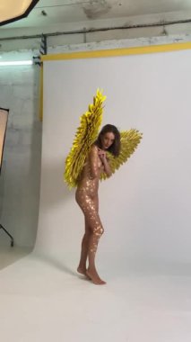 Altın kanatlı bir kadın stüdyoda duruyor, sahne arkasında altın boyayla kaplanmış çıplak bir vücut.