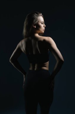 Bir kadın güçlü bir poz verir, silueti karanlık bir arkaplanda parlar. Yumuşak ışık, onun tanımlanmış özelliklerini ve zarif duruşunu vurguluyor..