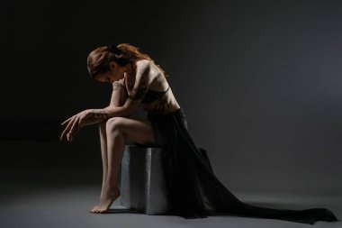 Dansçı bir tabureye oturur, aşağı bakar zarafetle, yumuşak ışık altında akan kumaşa sarınır..