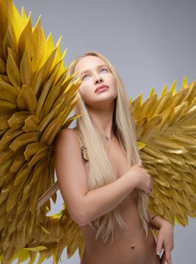 Uzun saçlı bir model zarif bir şekilde ayakta duruyor. Tarafsız bir zemine karşı nefes kesici altın kanatlar sergiliyor..