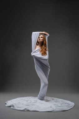 Zarif bir dansçı, yumuşak beyaz kumaşa sarılı halde sanatsal ifadeyi vurgulayarak akıcı hareketler sergiler..