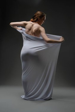 Bir kadın zarif bir şekilde kendini yumuşak kumaşa sarar, loş bir stüdyoda sanatsal hareketler sergiler..