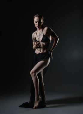 A model stands confidently in a dark studio, wearing a sleek outfit and showcasing intricate tattoos while interacting with flowing fabric. clipart