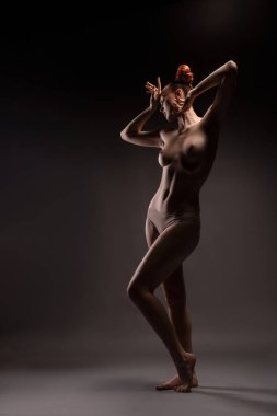 Çıplak göğüs ve külotlu kızıl saçlı zarif, genç bir bayan model. Gri arka planda gözleri kapalı bir yüze dokunuyor.