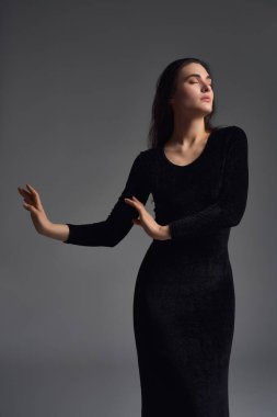 Zarif siyah elbiseli, kendine güvenen genç bir kadın. Kolunu kaldırıp gri arka planda kameraya bakıyor.