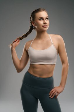 Genç bir kadın kendine güvenen bir ifadeyle duruyor, spor sutyen ve yüksek bel tayt giyiyor. Örgülü bir saç stili ve atletik bir duruşu var. Formda bir fiziği var..