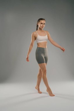 Kendine güvenen bir model pürüzsüz bir yüzey boyunca zarif adımlar atar, atletik bir giysi sergiler. Tarafsız arka plan onun duruşunu ve kıyafet tasarımını vurguluyor, fitness ve stili vurguluyor..