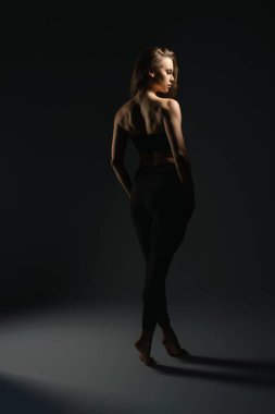 Karanlık bir stüdyoda yumuşak bir ışık altında zarif bir poz sergileyen bir model. Işık ve gölgenin etkileşimi onun siluetini çok güzel vurguluyor..