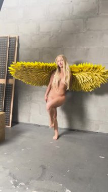 Bir kadın stüdyoda büyük sarı kanatlı üstsüz poz veriyor, eşsiz vücut sanatı ve yaratıcılığı sergiliyor..