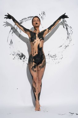 Sıska kadın siyah boyayla kaplanmış kameraya bakarken beyaz arka planda el izleriyle daireler çiziyor.