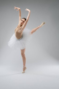 Bir balerin, bir performans sırasında esneklik ve sanatsallığı vurgulayarak, sade bir arka planda beyaz etekli zarif bir poz sergiler..