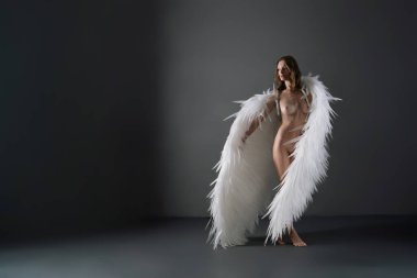 Çıplak bir kadın, arkasında büyük beyaz kanatları olan bir stüdyoda duruyor. Kanatlar vücudunun ötesine uzanarak çıplak poz vererek dramatik ve ruhani bir etki yaratıyor.