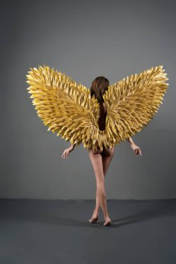 Altın kanatlı bir kadın kameradan uzaklaşır, arkası izleyiciye döner..