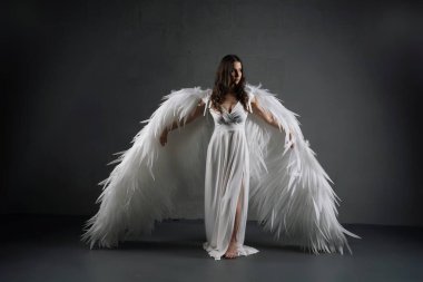 Bir kadın, arkasında büyük beyaz melek kanatlarıyla stüdyoda duruyor. Beyaz bir elbise giyiyor ve uzun siyah saçları var..