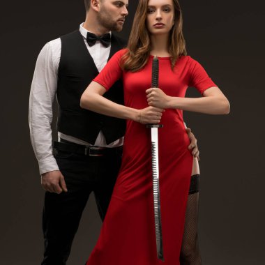 Stüdyoda şık bir çift yan yana durur, kırmızı elbiseli kadın kılıcını tutarken erkek arkasında kendinden emin bir poz verir..