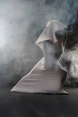 Örtülü bir figür dumanla etkileşime girer, akışkan hareketler gösterir ve sanatsal bir ortamda gizem uyandırır..