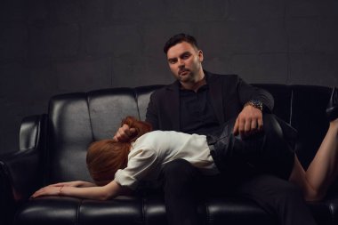 Düşünceli genç bir erkek kameraya bakıyor ve kız arkadaşının saçını tutuyor. Deri koltukta oturuyor ve karanlık stüdyoda kalçalarına dokunuyor.