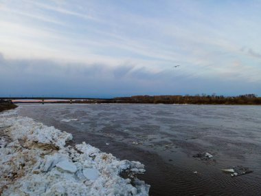 Sakin bir kış nehri manzarası buz oluşumlarını ve alacakaranlık altında bir köprüyü gösterir..