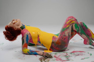 Tarafsız bir arkaplana yaslanan bir kadın, vücudu renkli boyalarla kaplı, yaratıcı bir gösteride sanat ve güzelliğin bir karışımını sergiliyor..