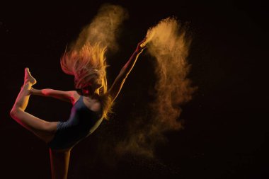 Canlı kıyafetli bir dansçı zarif bir şekilde zıplar, koyu bir arka plana doğru hareket ederken renkli tozlar gönderir..