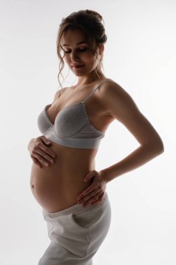 Güzel hamile bir kadın zarif bir şekilde duruyor, bebek karnındaki şişliği gösteren iç çamaşırları giyiyor, zarafet ve annelik duygusunu iletiyor..