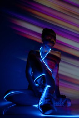 Bir kadın zarif bir şekilde oturur, çıplak bedeni bir projektörden gelen sanatsal ışık desenleriyle kaplıdır, güzelliğini ve zarafetini gösterir..