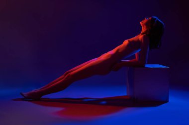 Mavi ve kırmızı neon ışıklarıyla aydınlatılmış, karanlık stüdyoda küpe yaslanan çıplak kadın modelinin tüm vücudu.