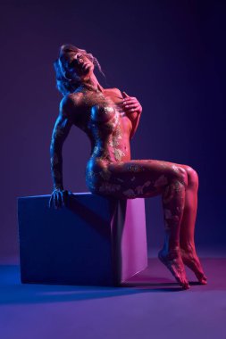 Neon ışıklarının altında, gözleri karanlık stüdyoda kapalı bir küpe yaslanmış, altın kaplamalı zarif çıplak kadının yan görüntüsü.