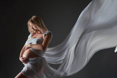 Uçan beyaz elbiseli hamile bir kadın göbeğini siyah arka planda kucaklıyor.