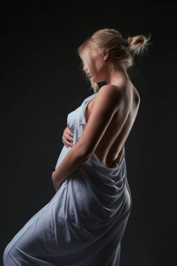 Şehvetli, anonim, hamile bir kadının, elleri karnında çıplak bir şekilde vücudunu saran zarif beyaz kumaşla aşağıya bakması.