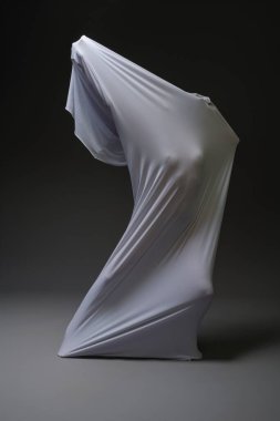 Beyaz kumaşa sarılmış bir figür, karanlık bir ortamda ışık ve gölge oyunlarını vurgulayan anlamlı bir duruş sergiliyor..