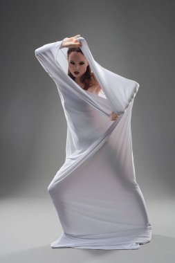 Beyaz bir elbiseye sarılı bir dansçı zarif hareketlerini bir stüdyo ortamında sergiliyor..