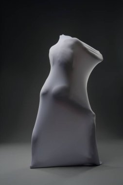 Örtülü bir figür pürüzsüz bir kumaştan çıkar ve karanlık bir arkaplana karşı soyut bir siluet yaratır..