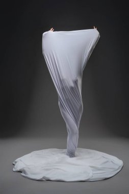 Bir insan sarmal beyaz bir kumaş içinde dramatik bir şekilde ortaya çıkar ve loş bir stüdyoda sanatsal bir görsel etki yaratır..